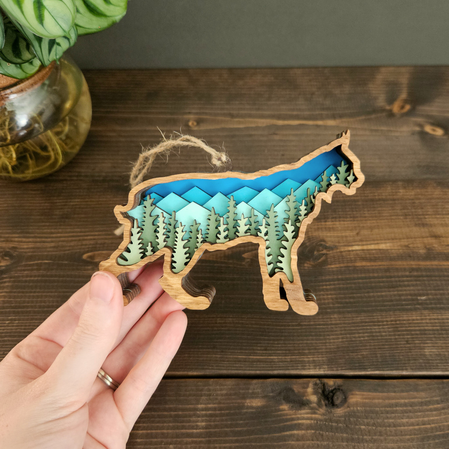 Wooden Christmas ornament shaped like a bobcat or lynx with a mountain and forest design held by a hand on a wooden surface.