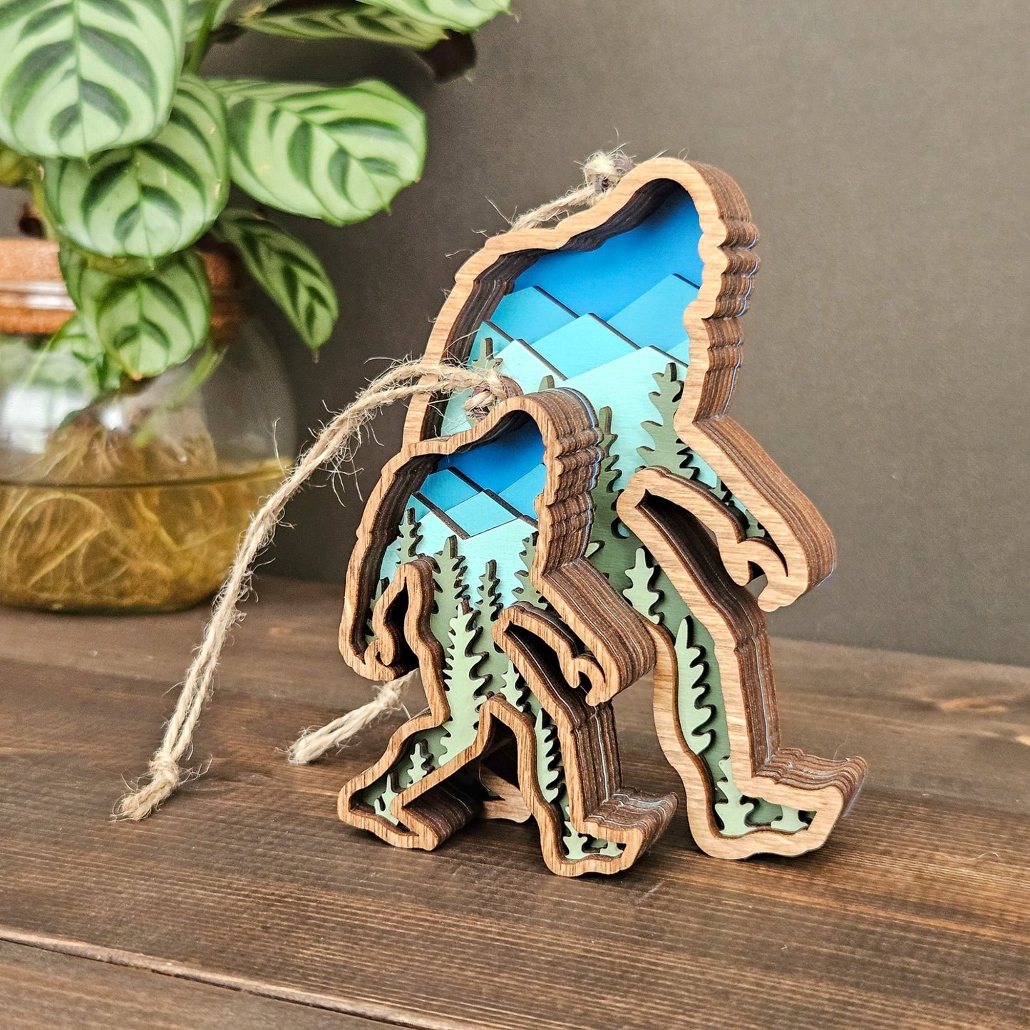 Wooden ornament shaped like a bigfoot with blue and green design, placed on a rock with a pine cone and moss.