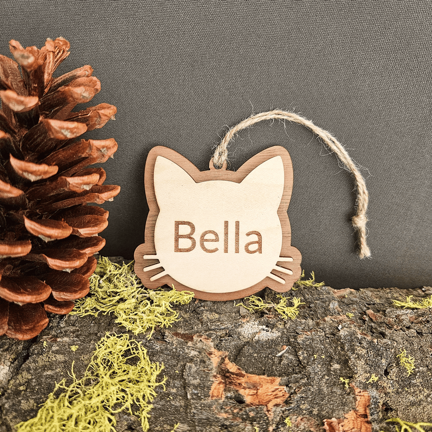 Personalized cat face ornament with name Bella displayed beside pinecone and moss on natural wood background