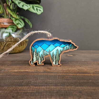 Decorative bear ornament with blue mountains and green tree design inside outline on a wooden surface