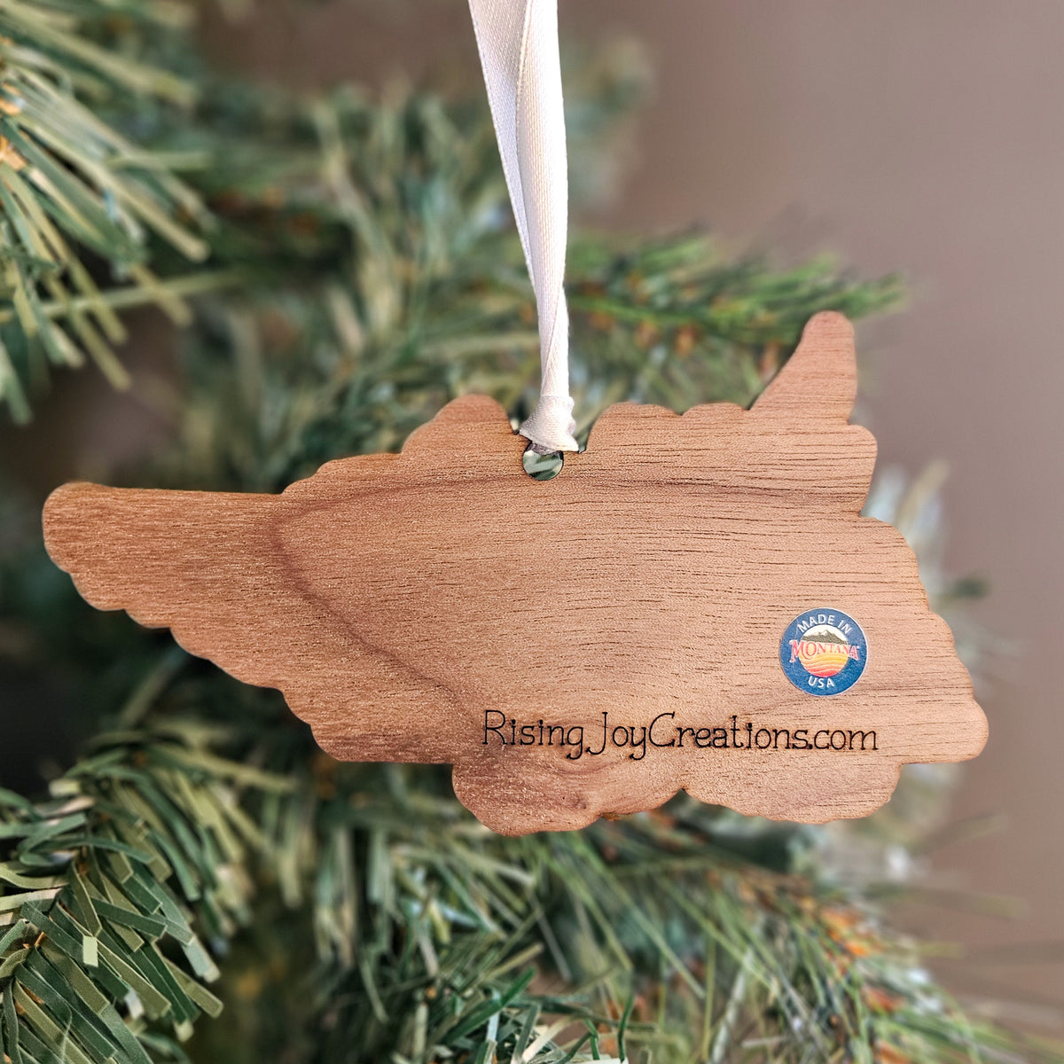 A wooden ornament shaped like the state of Texas hangs from a white ribbon on a green Christmas tree. The ornament has the text 'Rising Joy Creations.