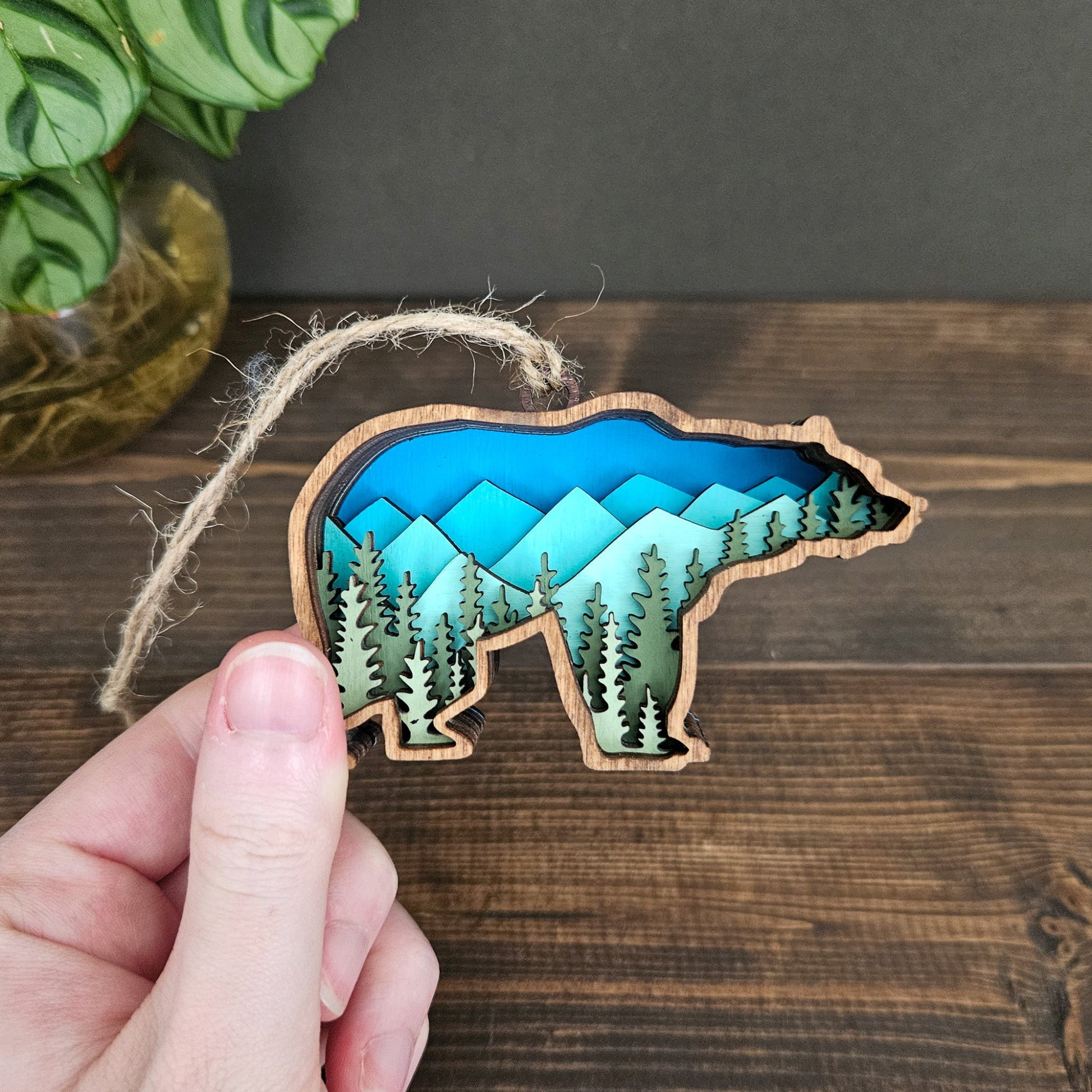Wooden bear-shaped ornament with mountain and forest design held by a hand on a wooden surface.