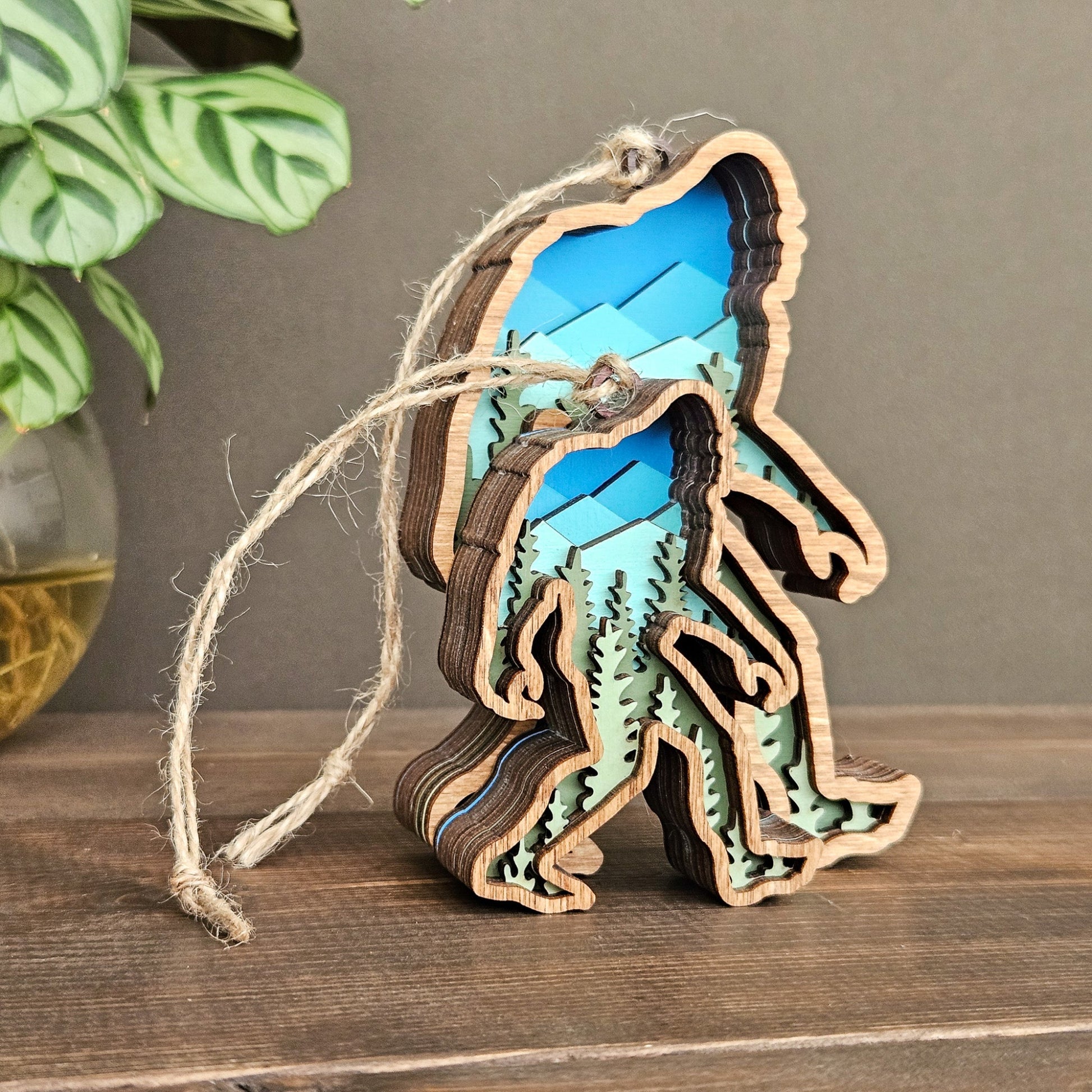 Wooden ornament shaped like a sasquatch with blue mountains and green trees