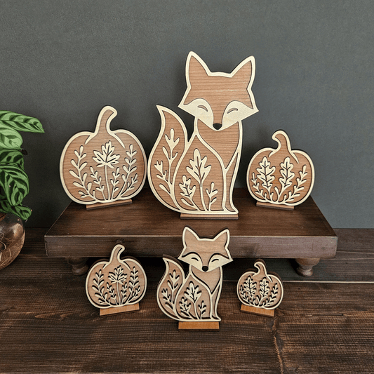 Wooden fox and pumpkin decorations on a wooden shelf with a plant in the background.