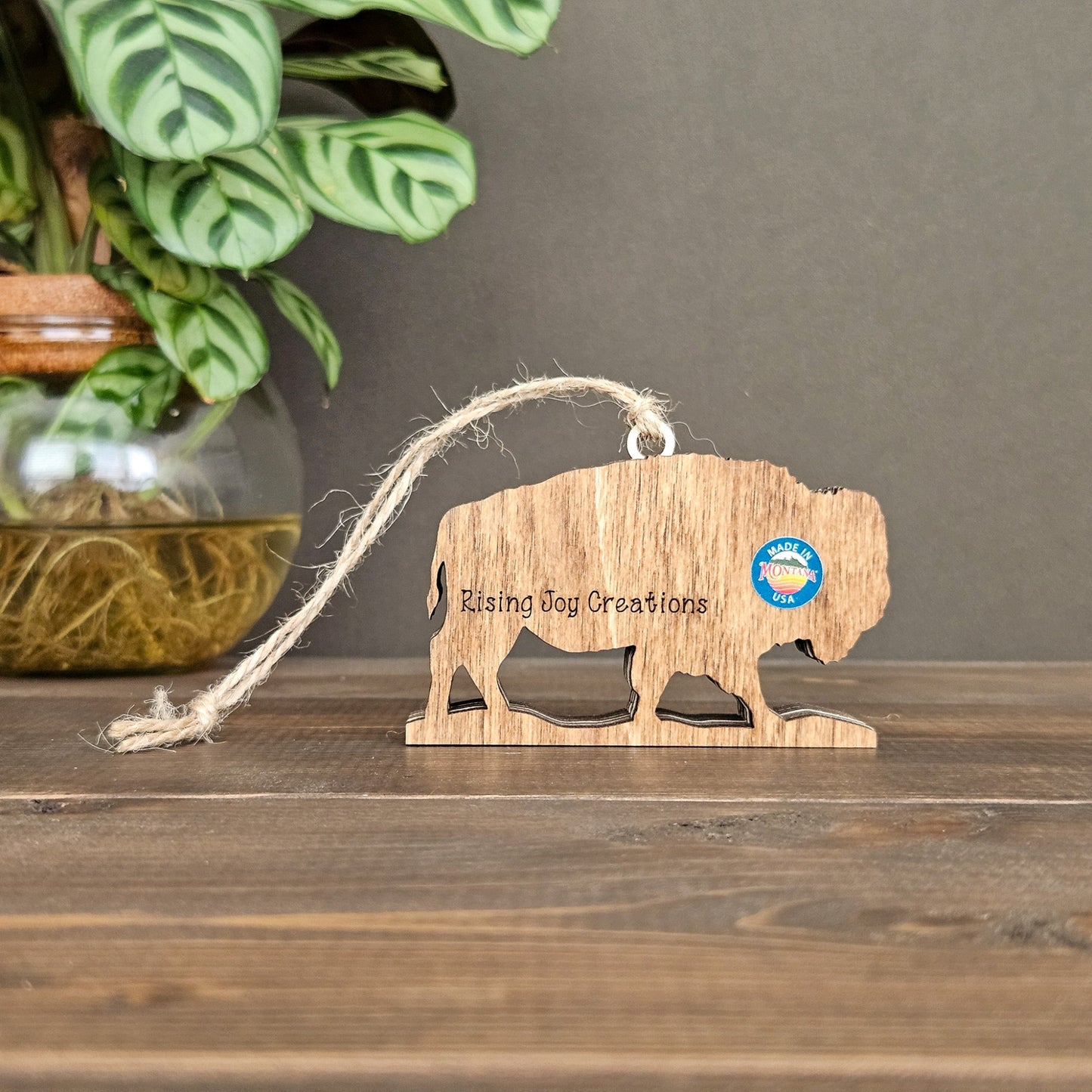 Wooden buffalo ornament with 'Rising Joy Creations' branding on a wooden surface with a plant in the background.