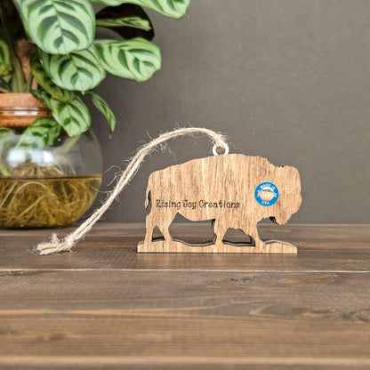 Wooden buffalo ornament with 'Rising Joy Creations' branding on a wooden surface with a plant in the background.