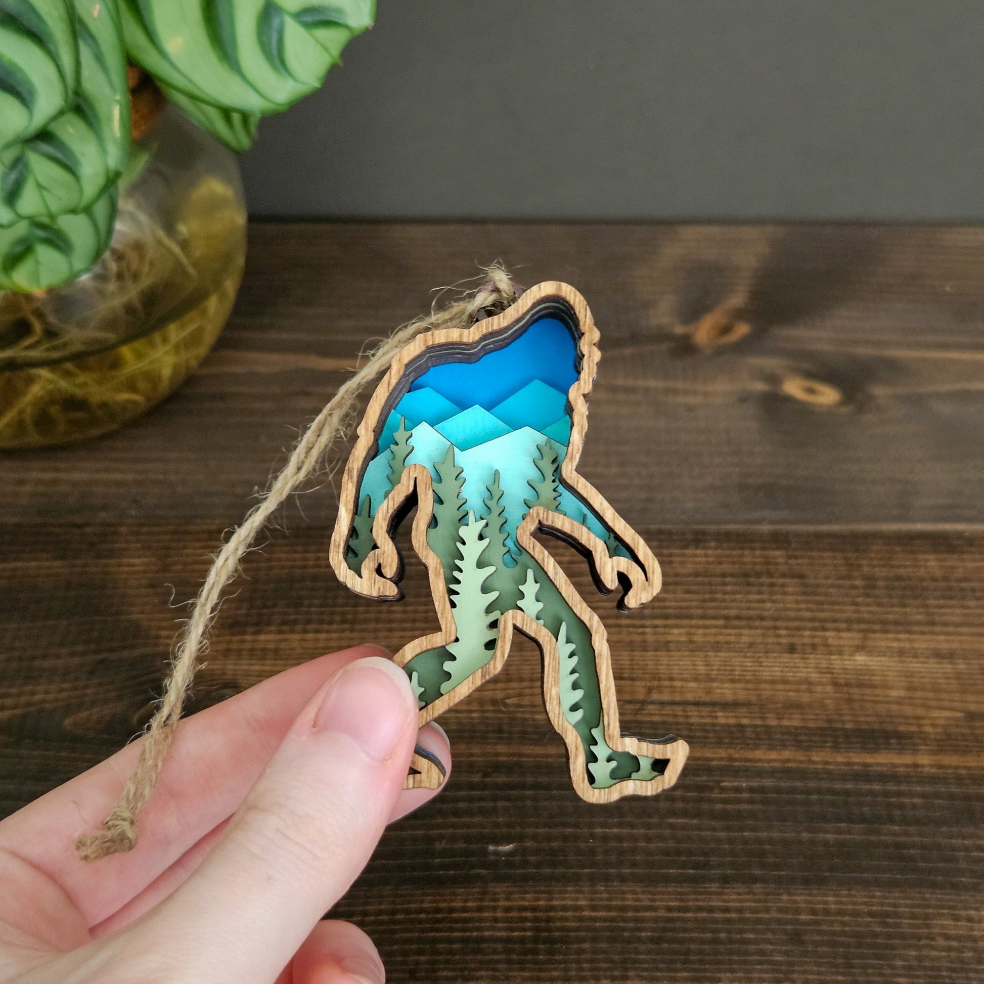 Hand holding a decorative ornament shaped like a bigfoot with a blue and green design on a wooden surface.