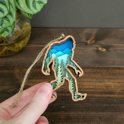 Hand holding a decorative ornament shaped like a bigfoot with a blue and green design on a wooden surface.