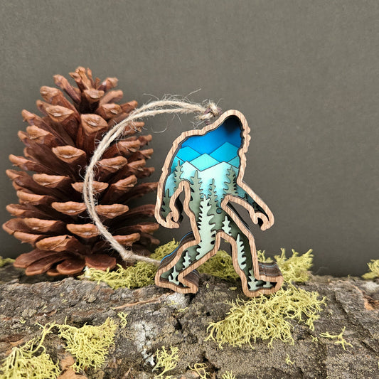 Wooden ornament shaped like a sasquatch with blue and green design, placed on a rock with a pine cone and moss.