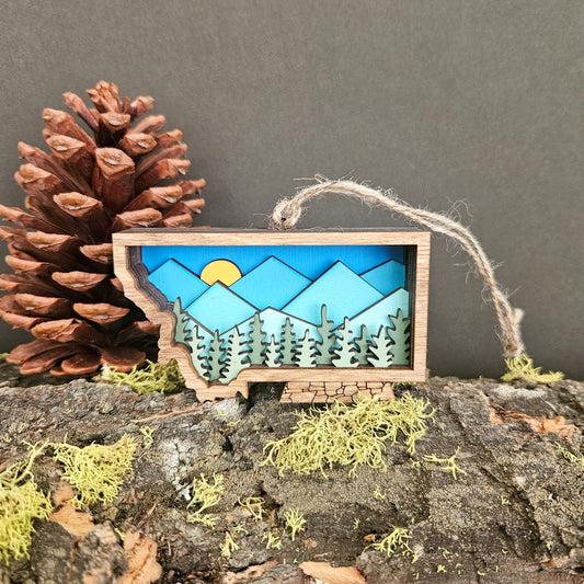 Wooden ornament with mountain and forest design on a rock with a pine cone and moss.