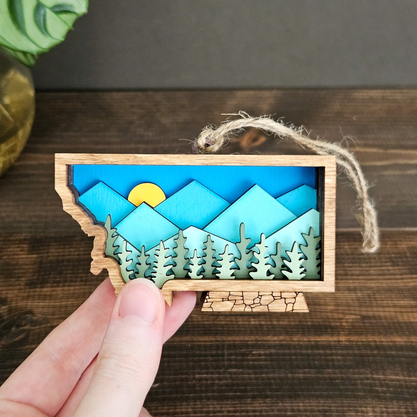 Wooden ornament with mountain and forest design held by a hand on a wooden surface.