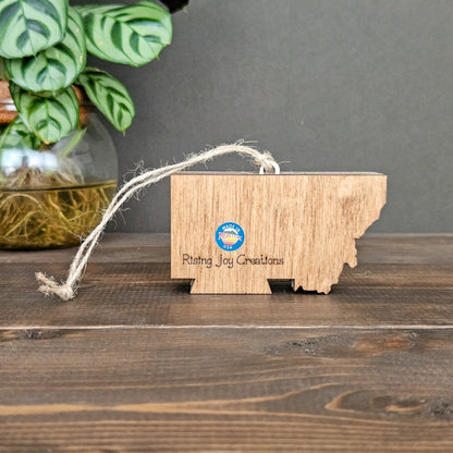 Wooden ornament shaped like a state with 'Rising Joy Creations' branding on a wooden surface.