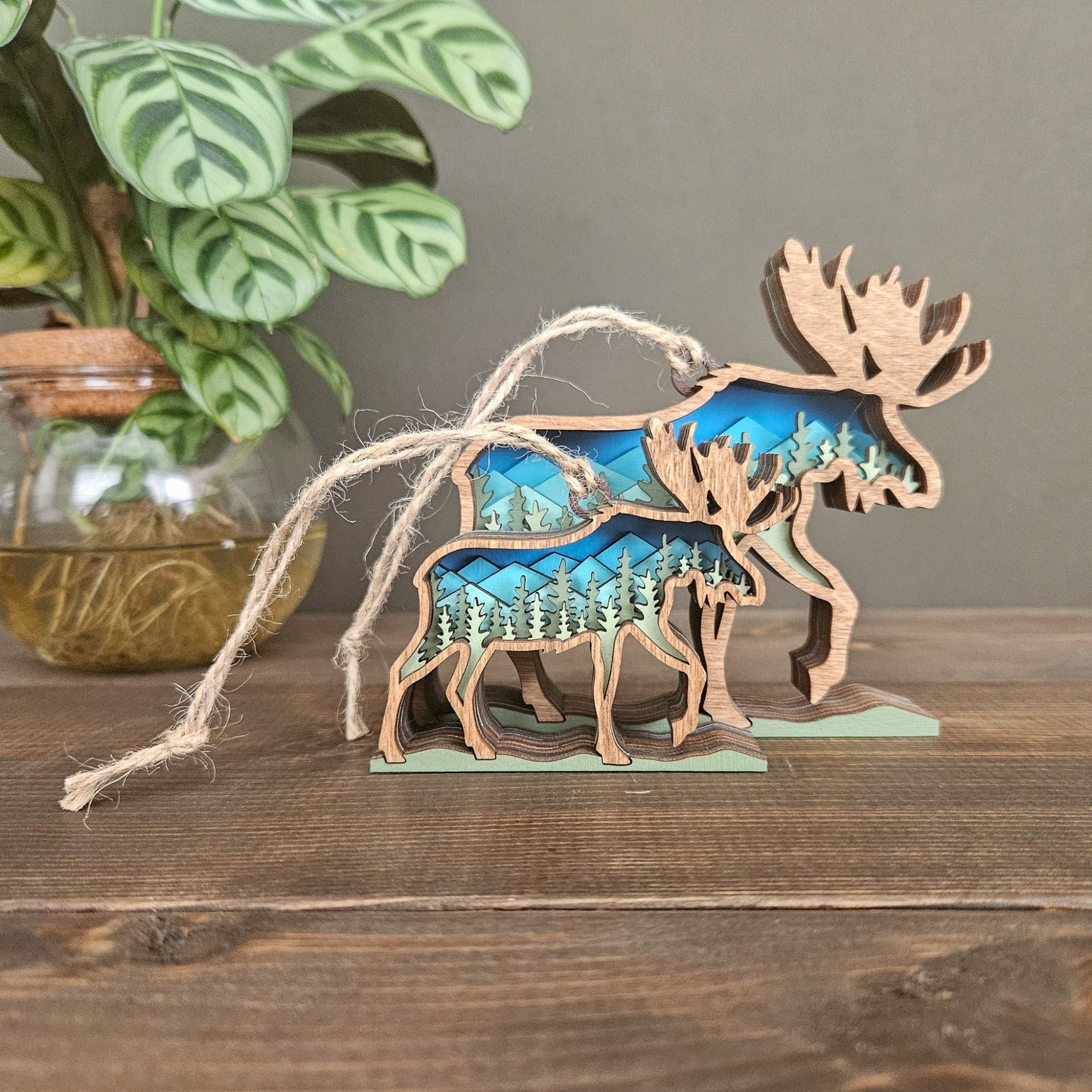 Wooden moose ornament with LED light inside, displaying a forest and mountain scene, on a wooden surface.