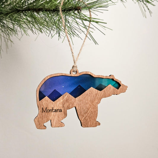 Wooden bear ornament with Norther Lights transparent material and woodmountain design and 'Montana' text, hanging on a tree.