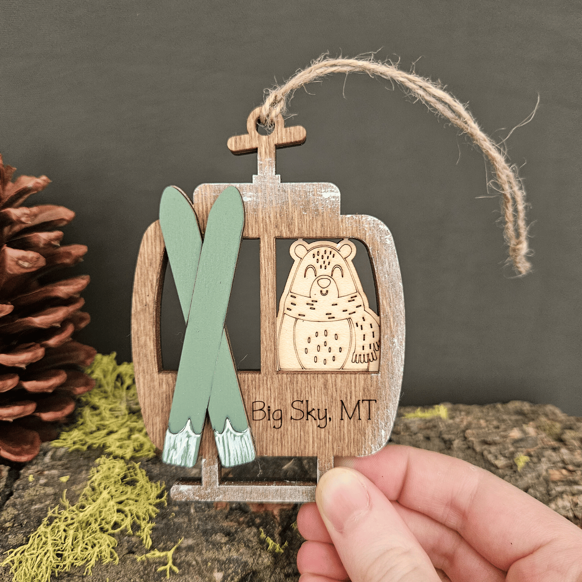 Wooden ornament with skis and bear design, held by a hand with a pine cone and greenery in the background.