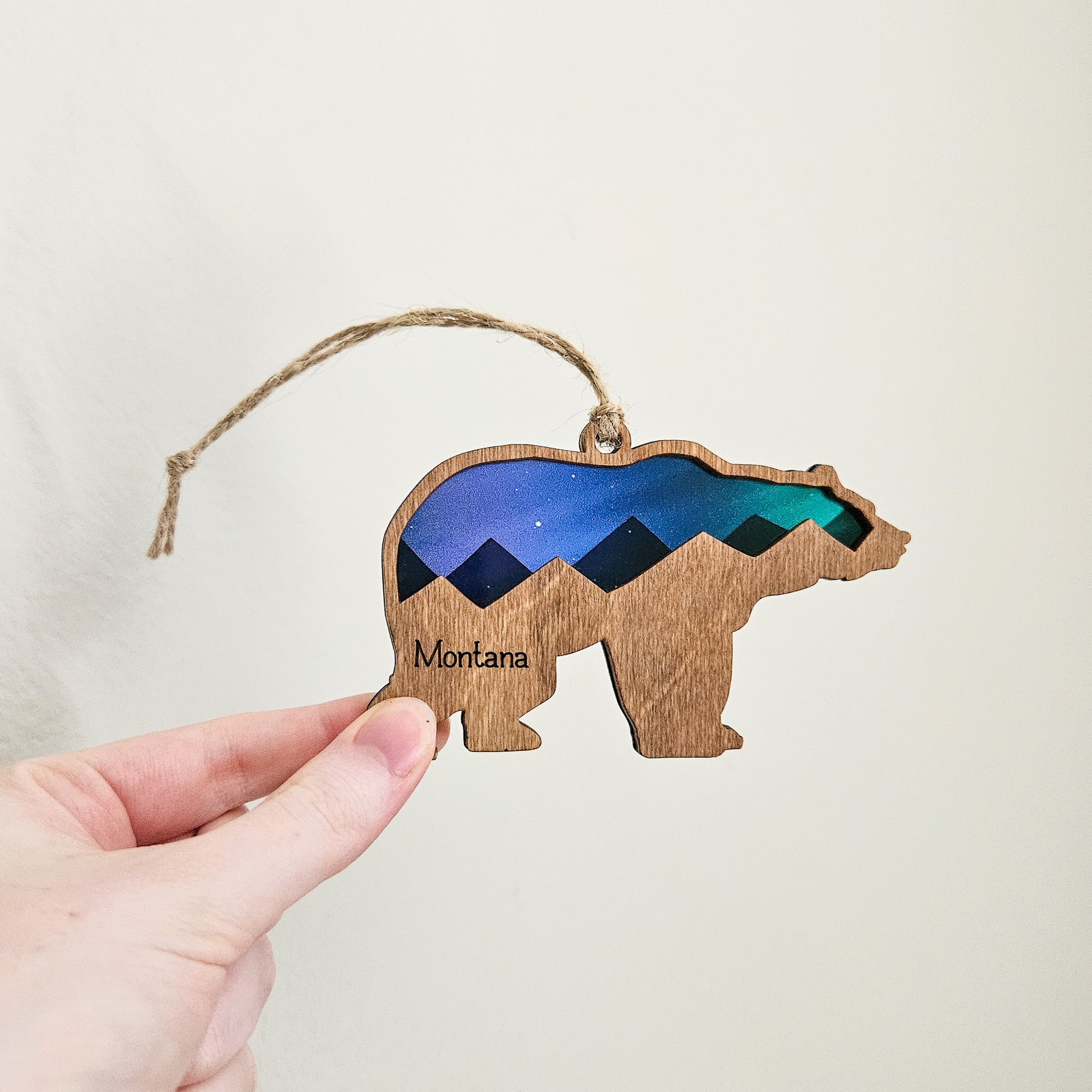 Wooden bear ornament with mountain design and 'Montana' text, held by a hand against a neutral background.
