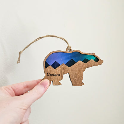 Wooden bear ornament with mountain design and 'Montana' text, held by a hand against a neutral background.