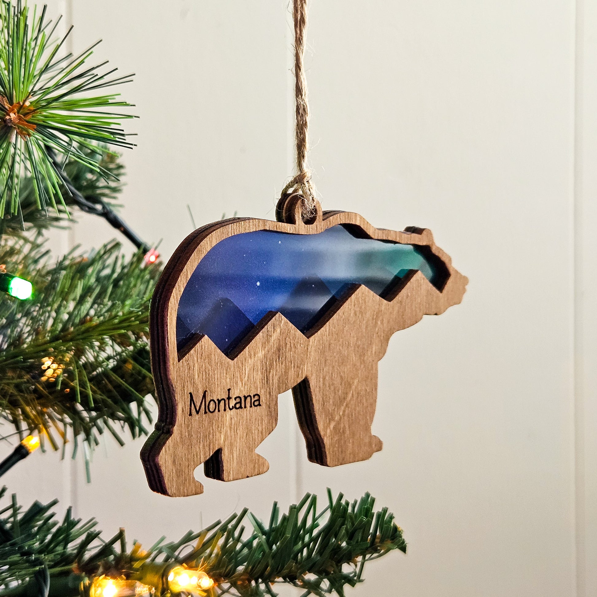 Wooden bear ornament with mountain design and 'Montana' text, hanging on a Christmas tree.