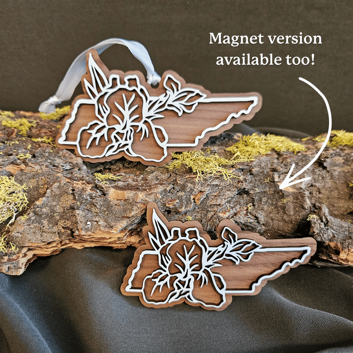 Wood Christmas ornament and magnet shaped like the state of Tennessee featuring a floral laser-cut design, displayed on a piece of mossy bark with a dark fabric backdrop. Ornament has a white hanging loop, and white text on the right reads 'Magnet version available too!' with an arrow pointing to the magnet version.