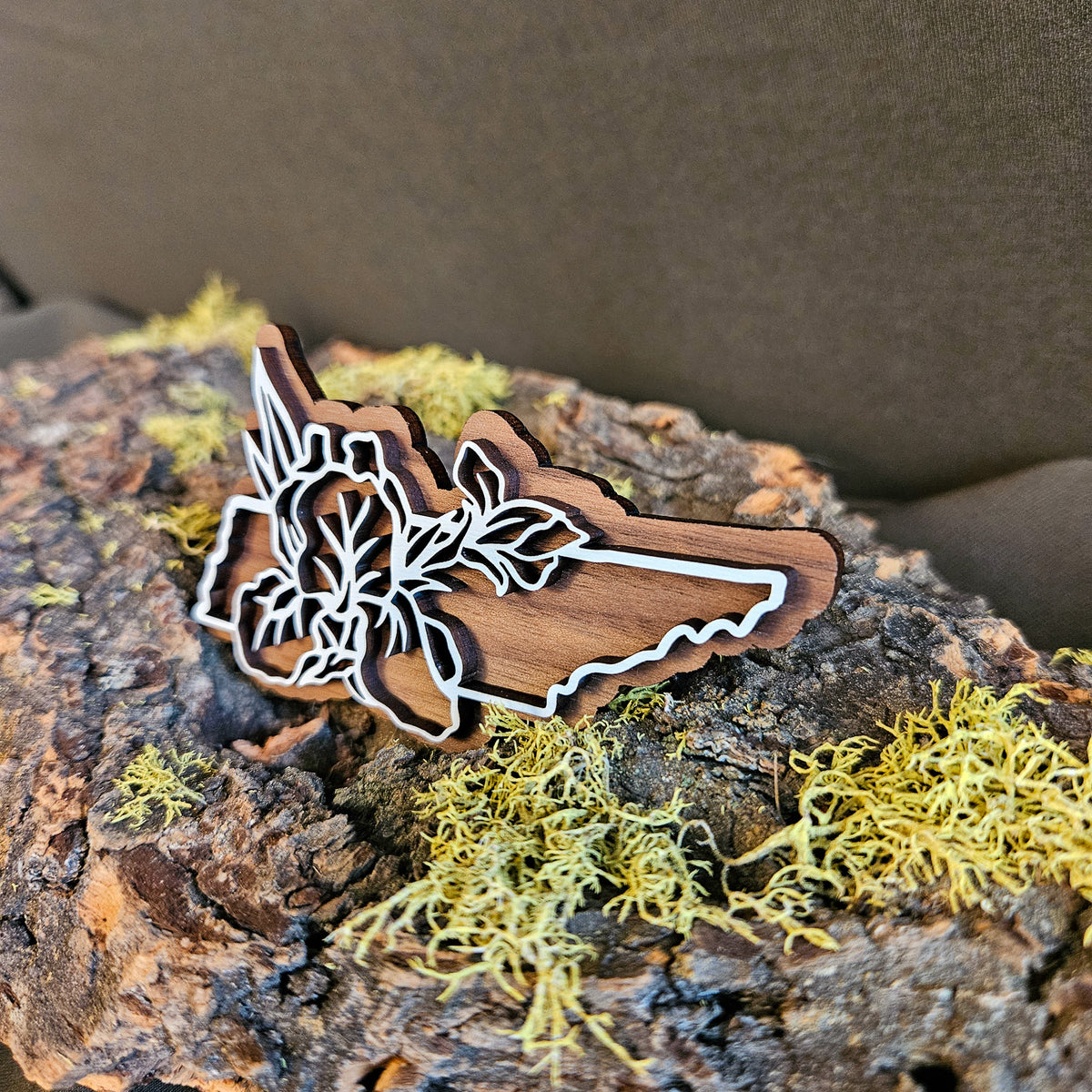A wooden cutout shaped like the state of Tennessee with white iris line art, displayed on textured bark with yellow-green moss against a dark brown background.