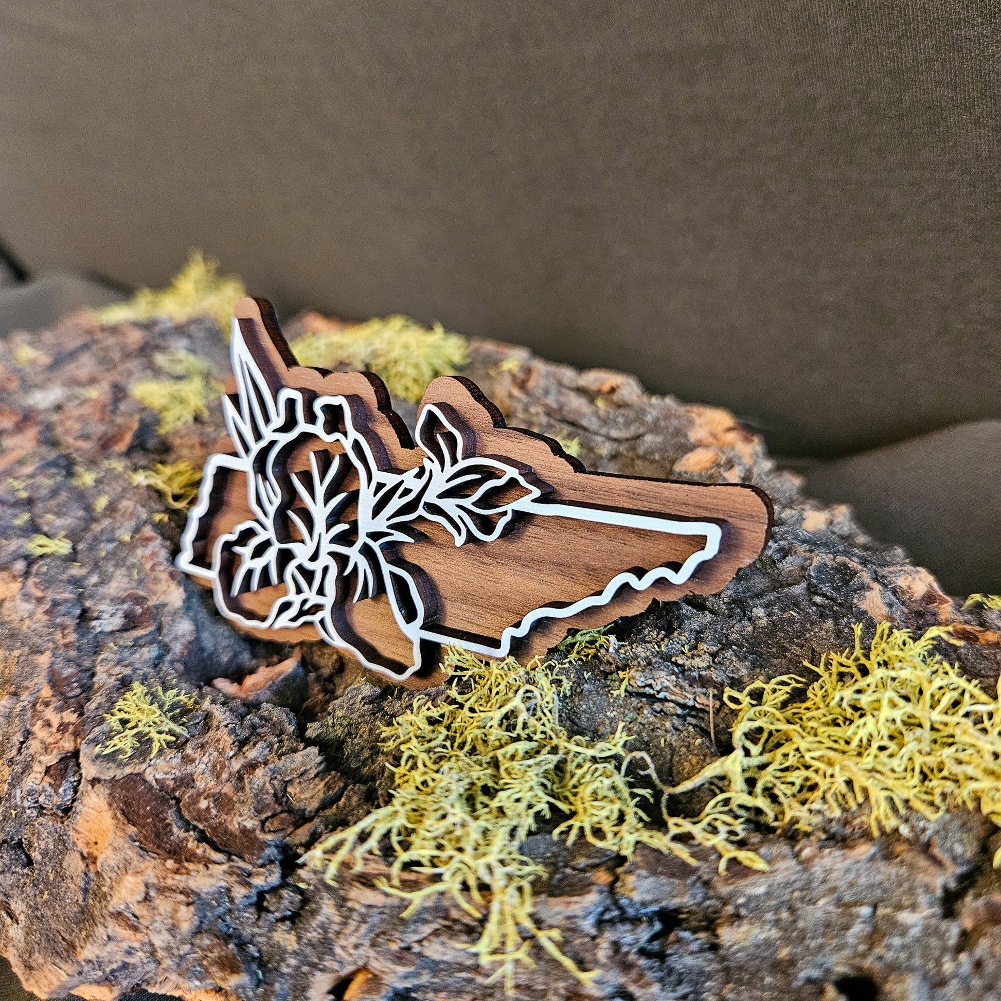 A wooden cutout shaped like the state of Tennessee with white iris line art, displayed on textured bark with yellow-green moss against a dark brown background.