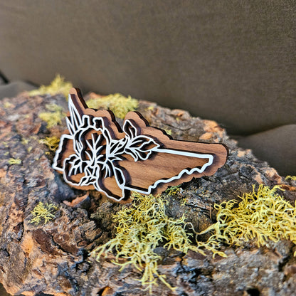 A wooden cutout shaped like the state of Tennessee with white iris line art, displayed on textured bark with yellow-green moss against a dark brown background.