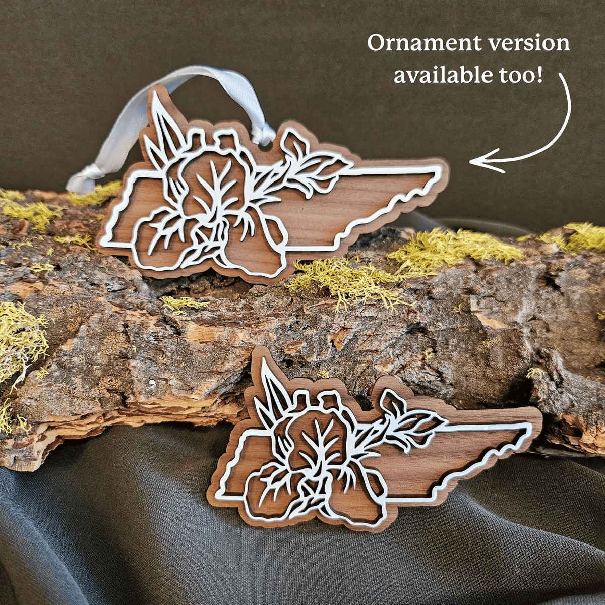 Wooden Tennessee-shaped ornament and magnet with white floral cutouts are displayed on a piece of bark with moss, with a dark fabric backdrop. Text in the upper right says 'Ornament version available too!' with an arrow pointing to one ornament.