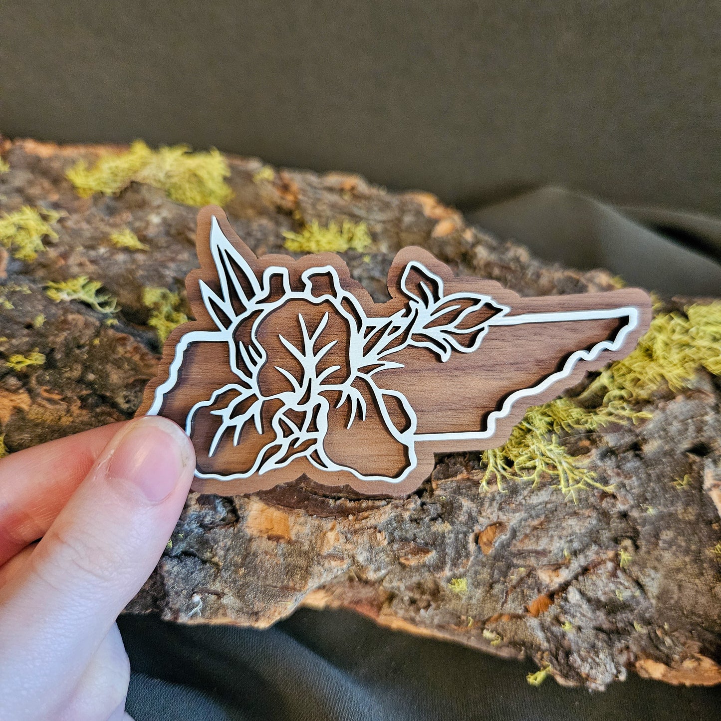 A hand holds a small wooden magnet shaped like the state of Tennessee, featuring a white laser-cut floral design over a natural wood-brown background, set against a piece of mossy bark.
