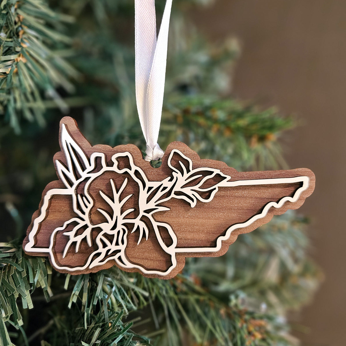 A wooden cutout Christmas ornament shaped like the state of Tennessee, featuring an Iris state flower design and hanging from a white ribbon, displayed on a green Christmas tree.