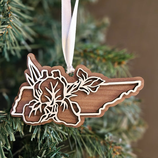 A wooden cutout Christmas ornament shaped like the state of Tennessee, featuring an Iris state flower design and hanging from a white ribbon, displayed on a green Christmas tree.
