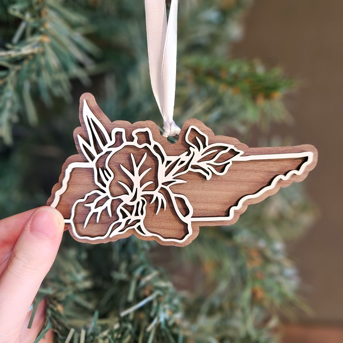 A hand holds a wooden Christmas ornament shaped like the state of Maine, featuring laser-cut floral and leaf designs, hanging from a cream ribbon against a blurred Christmas tree background.
