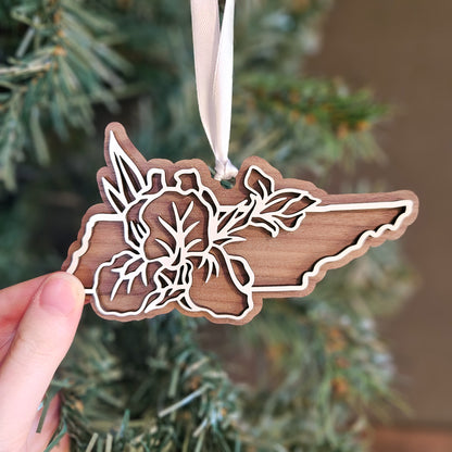A hand holds a wooden Christmas ornament shaped like the state of Maine, featuring laser-cut floral and leaf designs, hanging from a cream ribbon against a blurred Christmas tree background.