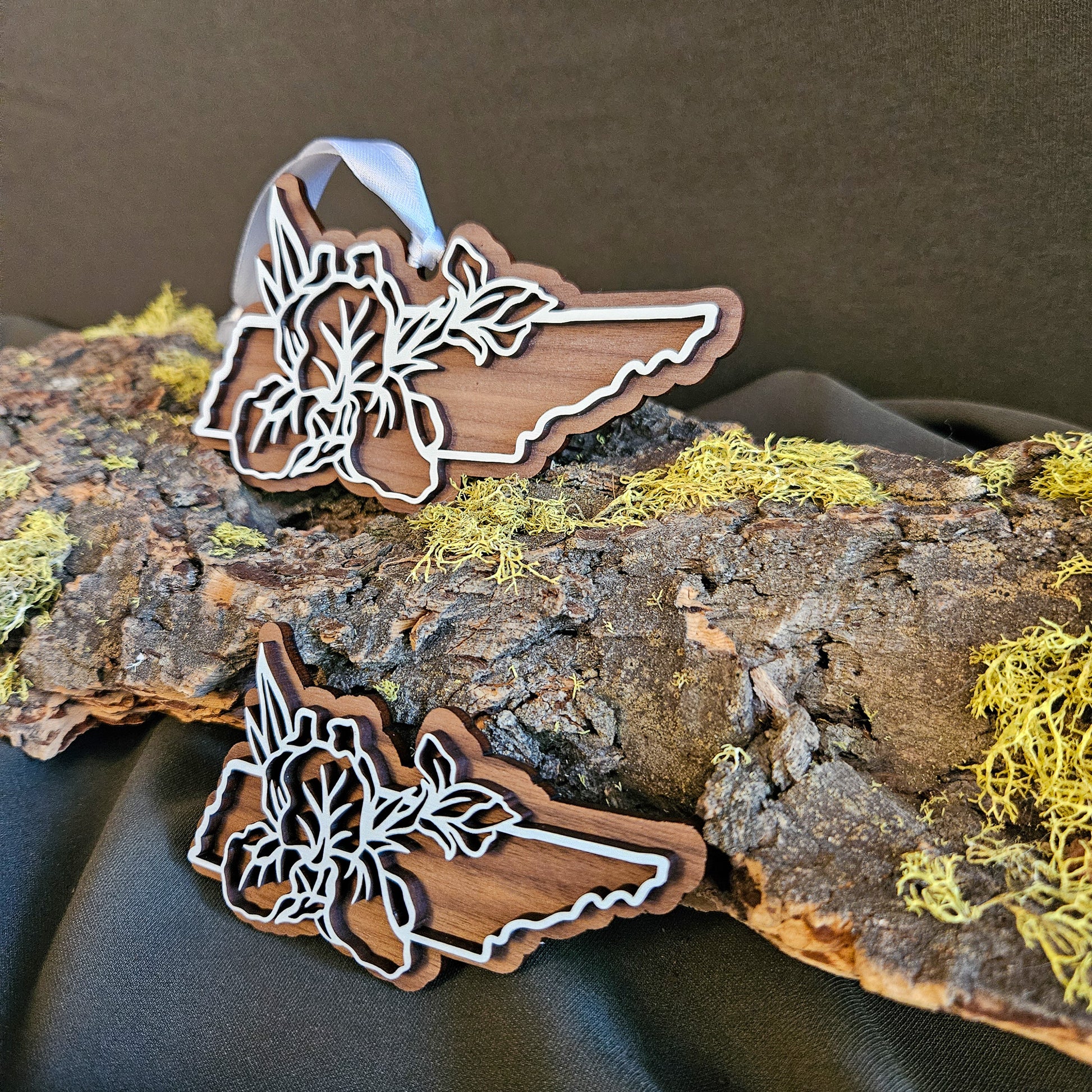 Two wooden cutout shape of Tennessee outline with floral designs, displayed on a piece of bark with yellow moss against a dark fabric background.