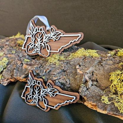 Two wooden cutout shape of Tennessee outline with floral designs, displayed on a piece of bark with yellow moss against a dark fabric background.