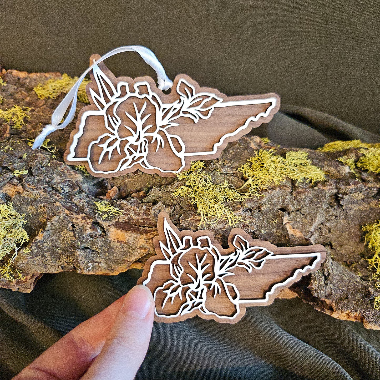 Two wooden Tennessee shapes with laser-cut floral design of the Iris, the state flower,  are displayed on a piece of bark with moss, with the magnet being held by hand.