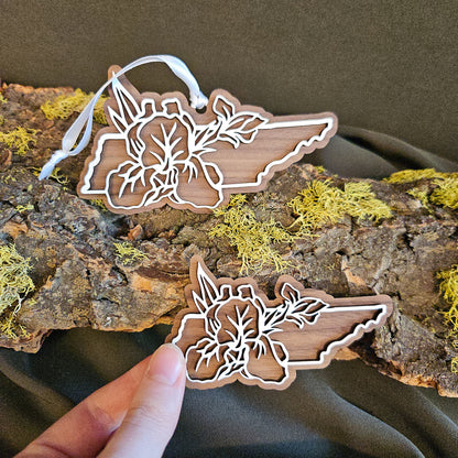 Two wooden Tennessee shapes with laser-cut floral design of the Iris, the state flower,  are displayed on a piece of bark with moss, with the magnet being held by hand.