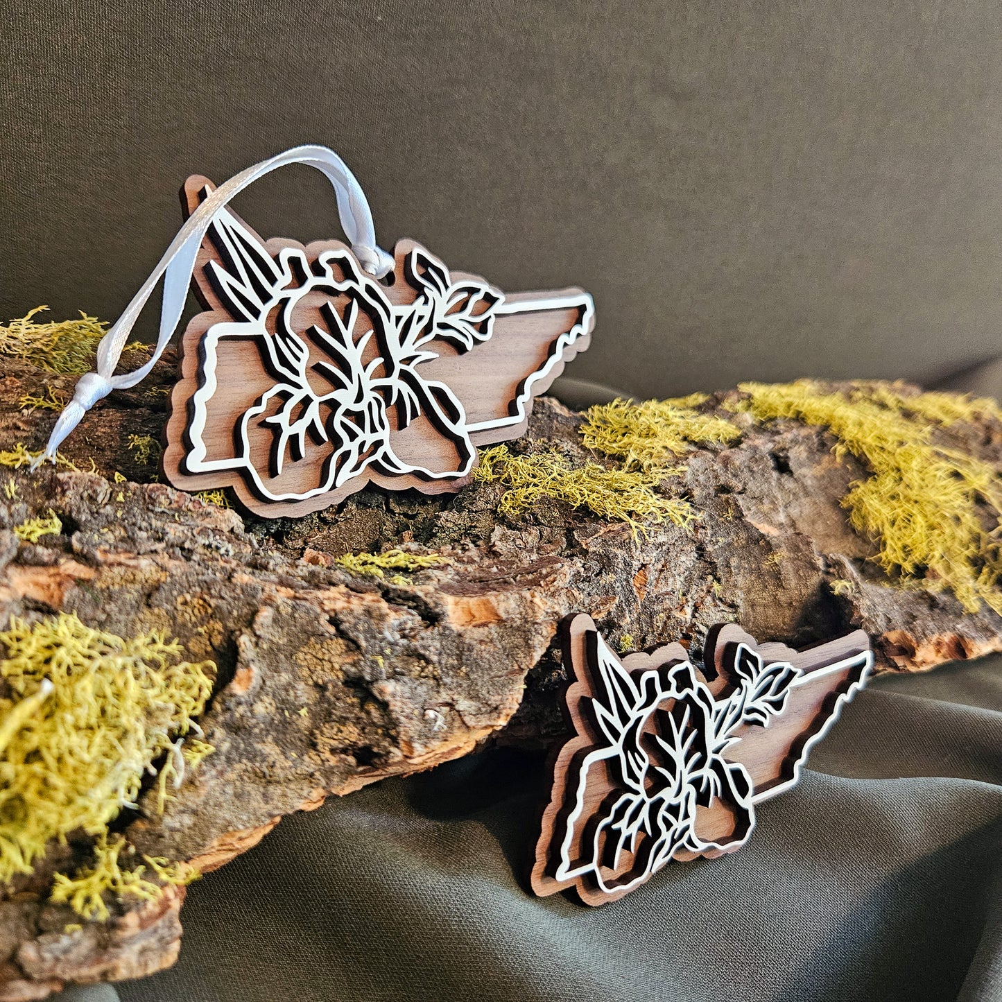 Wooden Christmas ornament and magnet shaped like Tennessee outline with a floral design, displayed on a mossy log against a dark fabric background.