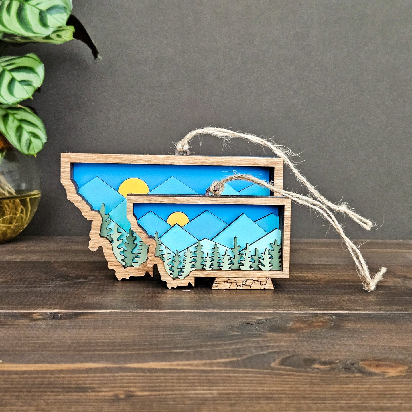 Wooden ornament shaped like a state with a mountain and sun design, hanging on a dark surface.