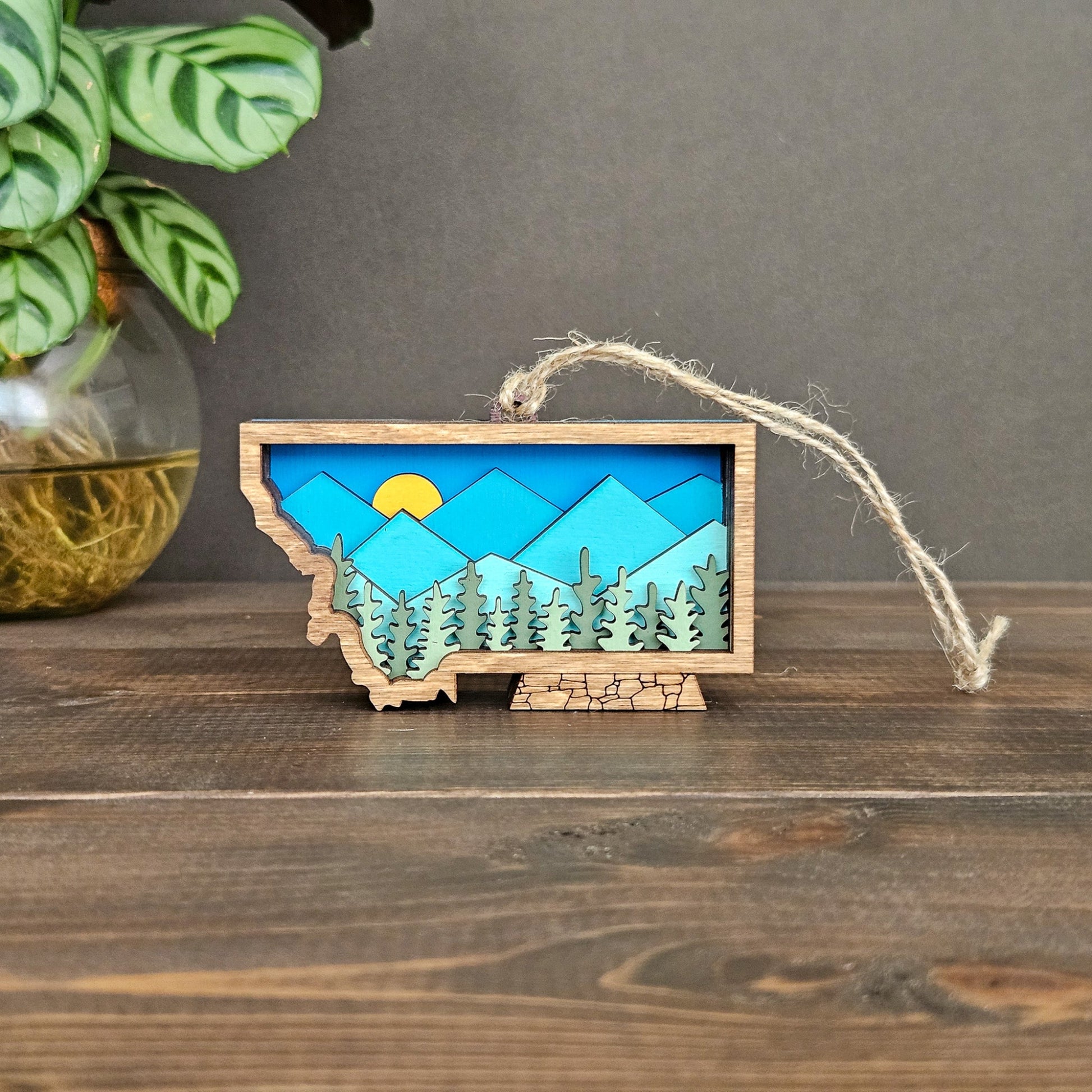 Decorative ornament shaped like the state of Montana with a mountain and sun design, hanging on a wooden surface.