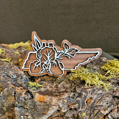 A wooden cutout shaped like the state of Tennessee, featuring a white laser-engraved floral design over walnut, displayed on a textured log with green moss against a dark brown background.