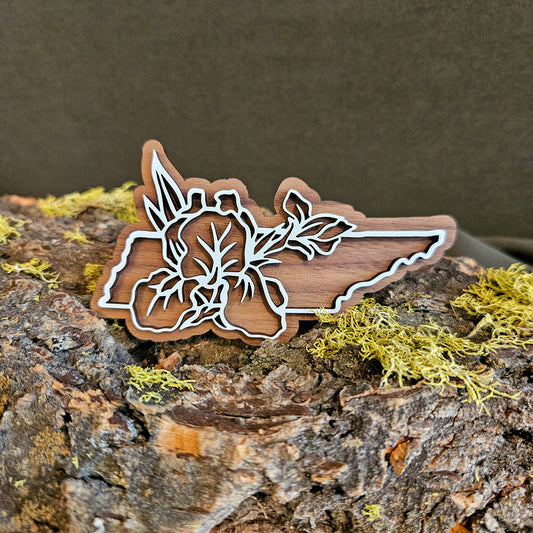 A wooden cutout shaped like the state of Tennessee, featuring a white laser-engraved floral design over walnut, displayed on a textured log with green moss against a dark brown background.