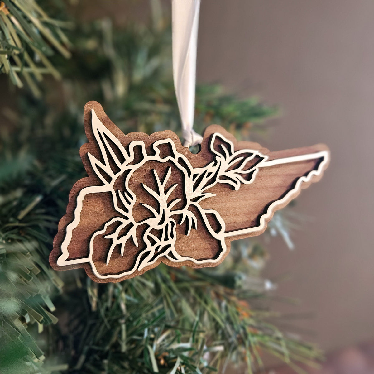 A wooden Christmas ornament shaped like the state of Tennessee with a floral laser-cut design, hanging from a white ribbon on a Christmas tree branch.