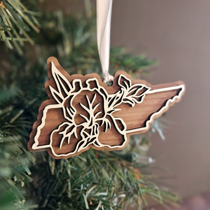 A wooden Christmas ornament shaped like the state of Tennessee with a floral laser-cut design, hanging from a white ribbon on a Christmas tree branch.