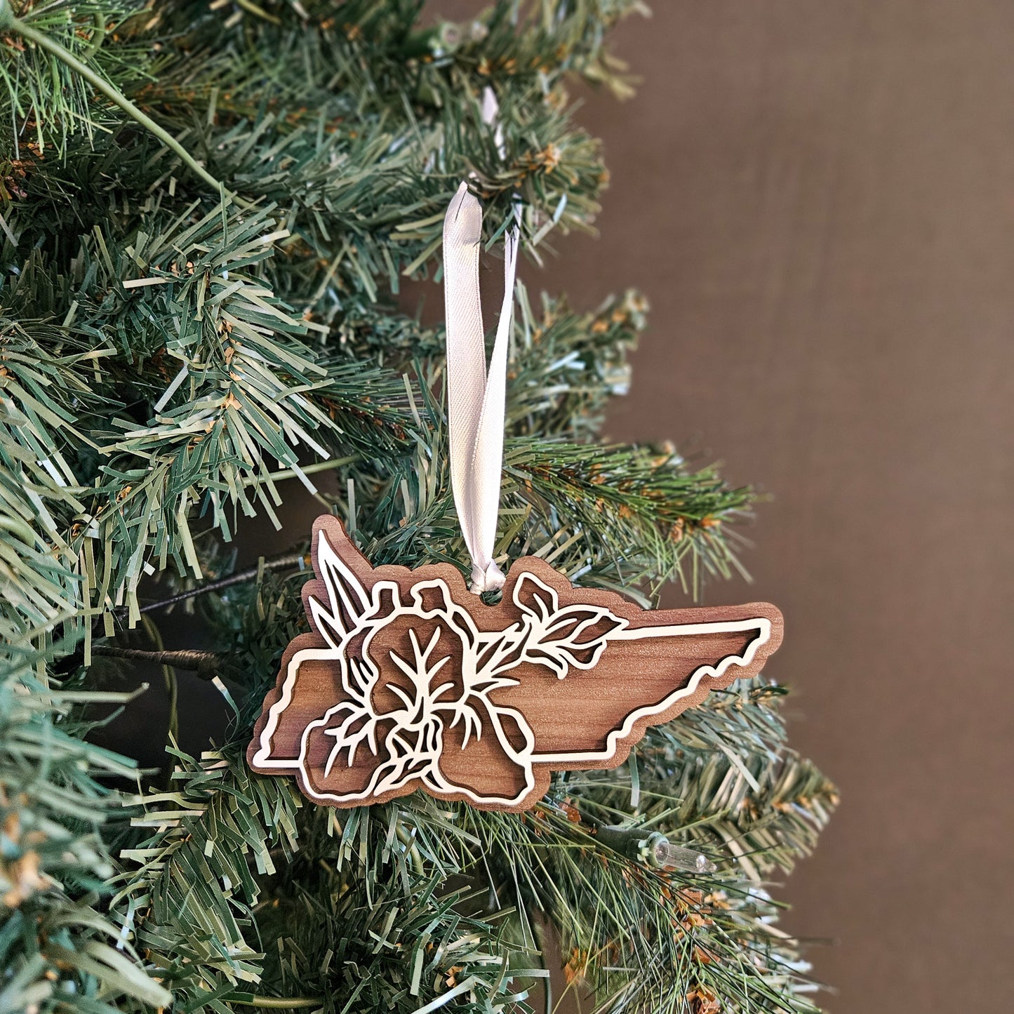 A wooden cutout Christmas ornament shaped like the state of Tennessee, featuring a floral design, hanging from a white ribbon on a green Christmas tree against a brown background.