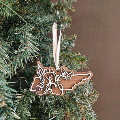 A wooden cutout Christmas ornament shaped like the state of Tennessee, featuring a floral design, hanging from a white ribbon on a green Christmas tree against a brown background.