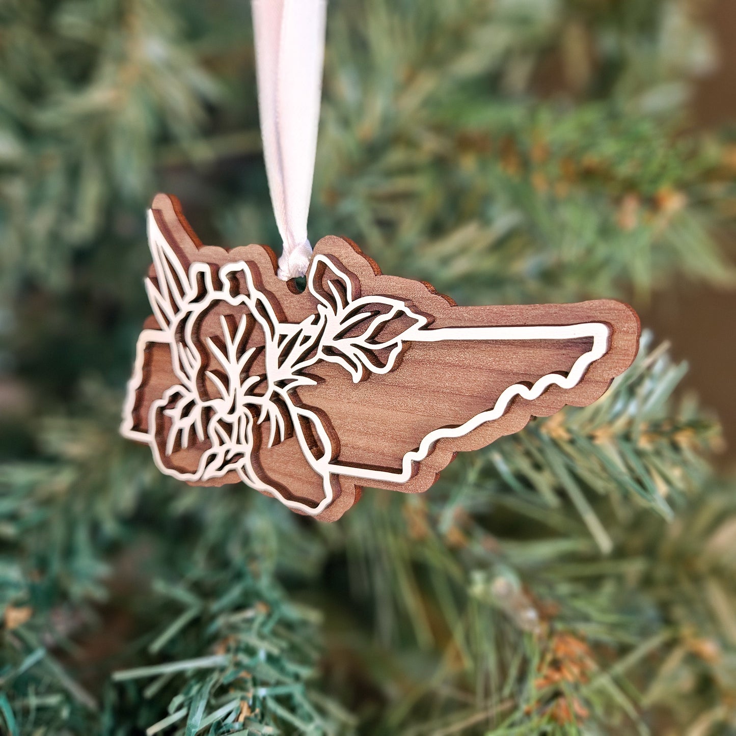 A wooden Christmas ornament shaped like the state of Rhode Island, featuring laser-cut floral details along the border, hanging from a white ribbon against a blurred green Christmas tree background.