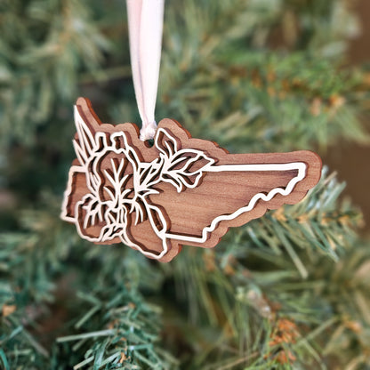 A wooden Christmas ornament shaped like the state of Rhode Island, featuring laser-cut floral details along the border, hanging from a white ribbon against a blurred green Christmas tree background.