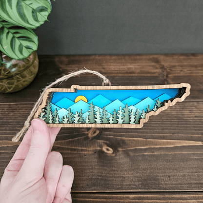 Hand holding layered Tennessee mountain ornament — handcrafted wooden state keepsake with sunrise and forest scene.
