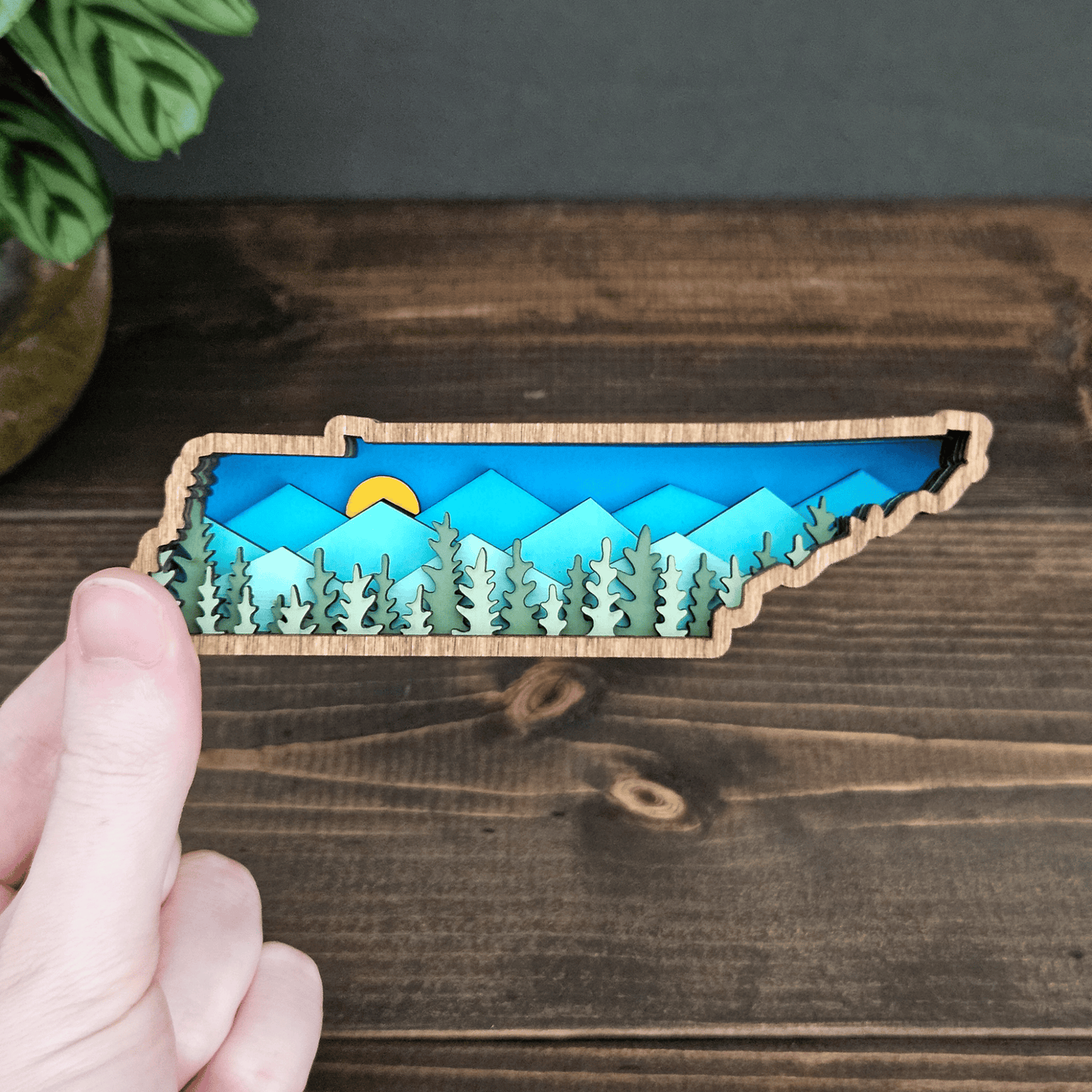 Hand holding mini layered Tennessee mountain magnet with forest and sunrise design — handcrafted wood state art by Rising Joy Creations.