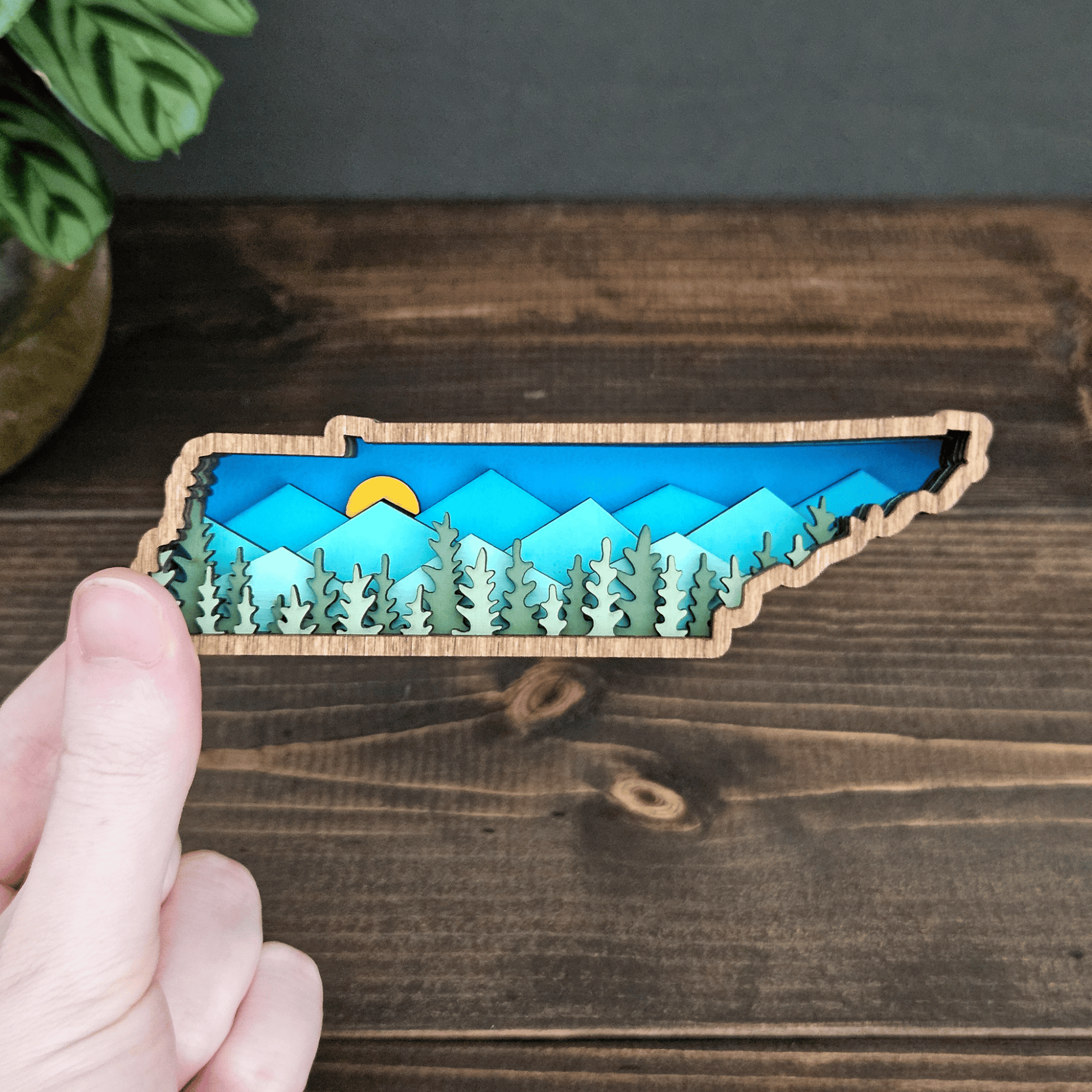 Hand holding mini layered Tennessee mountain magnet with forest and sunrise design — handcrafted wood state art by Rising Joy Creations.